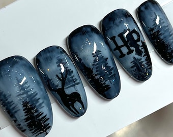 Puede incluir: Conjunto de cinco uñas postizas grises y negras con una escena de bosque y purpurina. Una uña presenta el logotipo de Harry Potter "HP" y otra tiene una silueta de ciervo.