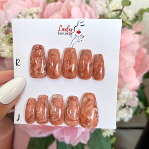 May include: Set of ten false nails with a brown and white marbled design. The nails are in a coffin shape and are arranged in two rows on a white card with the text "Lady Trousse" printed on it.