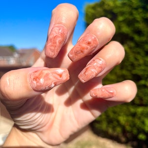 May include: Close-up of a hand with long, almond-shaped nails painted with a light peach and white marbled design.