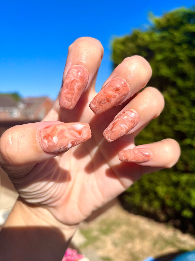 May include: Close-up of a hand with long, almond-shaped nails painted with a light peach and white marbled design.