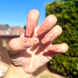 May include: Close-up of a hand with long, almond-shaped nails painted with a marbled design in shades of peach and white.