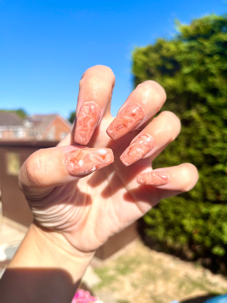 May include: Close-up of a hand with long, almond-shaped nails painted with a marbled design in shades of peach and white.