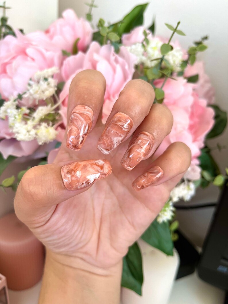 May include: Close-up of a hand wearing a set of peach-colored press-on nails with a white marble design.