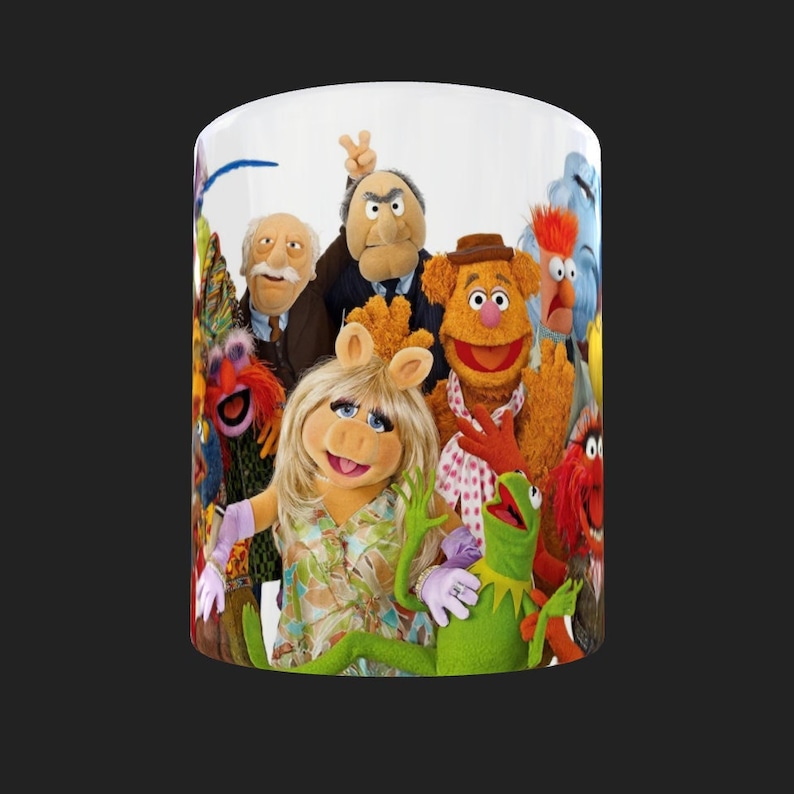 K&ouml;nnte beinhalten: Eine wei&szlig;e Keramiktasse mit einem bunten Bild der Muppet-Charaktere. Zu den Charakteren geh&ouml;ren Miss Piggy, Kermit der Frosch, Fozzie B&auml;r, Gonzo, Animal und andere.