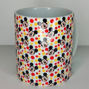 K&ouml;nnte beinhalten: Wei&szlig;e Keramiktasse mit einem sich wiederholenden Muster von Mickey-Maus-K&ouml;pfen auf wei&szlig;em Hintergrund mit roten und gelben Punkten.