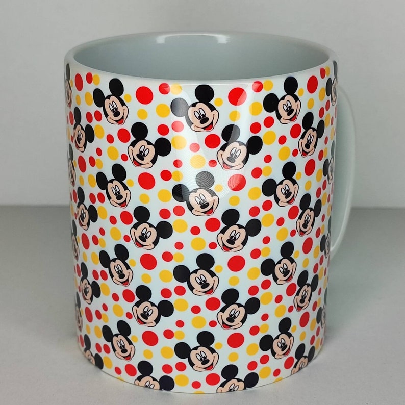 K&ouml;nnte beinhalten: Wei&szlig;e Keramiktasse mit einem sich wiederholenden Muster von Mickey-Maus-K&ouml;pfen auf wei&szlig;em Hintergrund mit roten und gelben Punkten.
