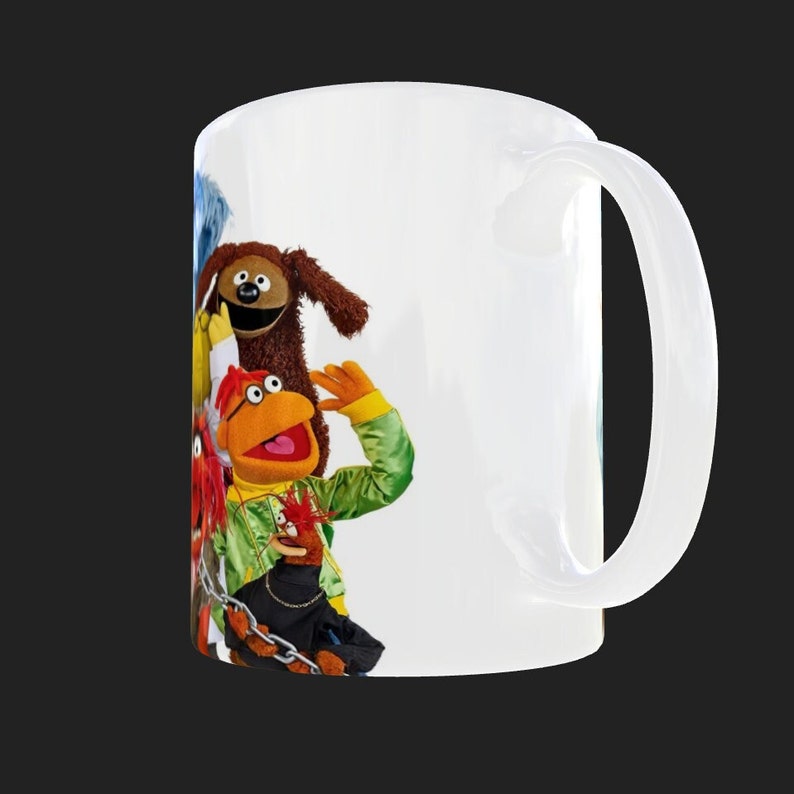 K&ouml;nnte beinhalten: Wei&szlig;e Keramiktasse mit einer bunten Illustration von vier Muppet-Charakteren. Die Charaktere sind ein blaues Monster, ein brauner Hund, ein roter Hahn und ein gelber Vogel. Die Tasse hat einen wei&szlig;en Henkel.
