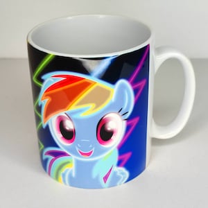 Könnte beinhalten: Eine weiße Keramiktasse mit einem neongelben und pinkfarbenen Design mit Rainbow Dash von My Little Pony. Der Hintergrund ist schwarz mit neongelben Blitzen.