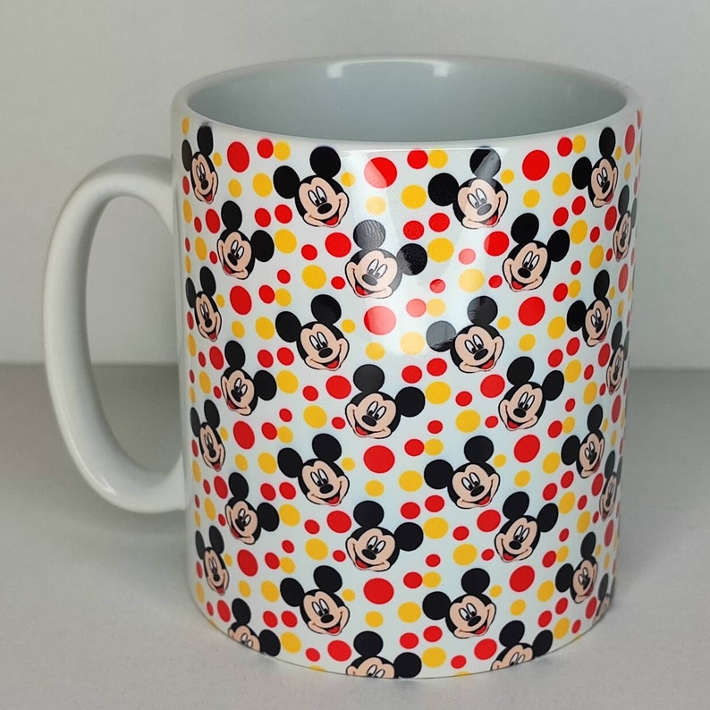 K&ouml;nnte beinhalten: Wei&szlig;e Keramiktasse mit einem sich wiederholenden Muster aus Mickey-Maus-K&ouml;pfen und roten und gelben Punkten.