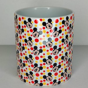 K&ouml;nnte beinhalten: Wei&szlig;e Keramiktasse mit einem sich wiederholenden Muster von Mickey-Maus-K&ouml;pfen auf wei&szlig;em Hintergrund mit roten und gelben Punkten.