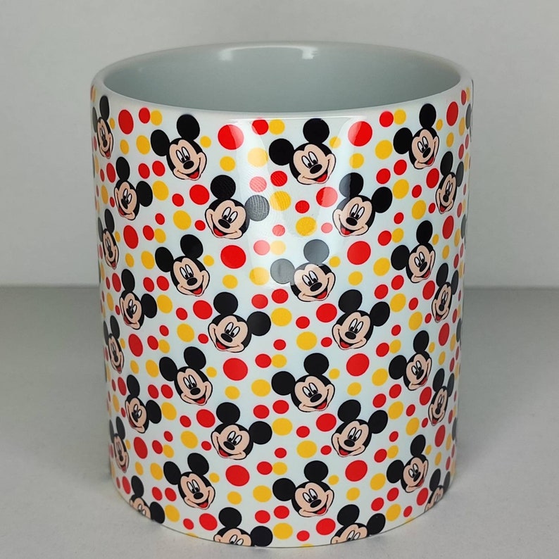 K&ouml;nnte beinhalten: Wei&szlig;e Keramiktasse mit einem sich wiederholenden Muster von Mickey-Maus-K&ouml;pfen auf wei&szlig;em Hintergrund mit roten und gelben Punkten.