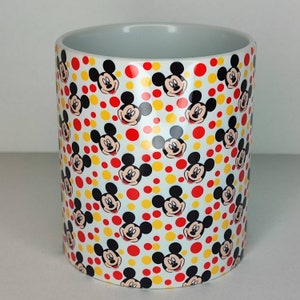 K&ouml;nnte beinhalten: Wei&szlig;e Keramiktasse mit einem sich wiederholenden Muster aus Mickey-Maus-K&ouml;pfen und gelben und roten Punkten.