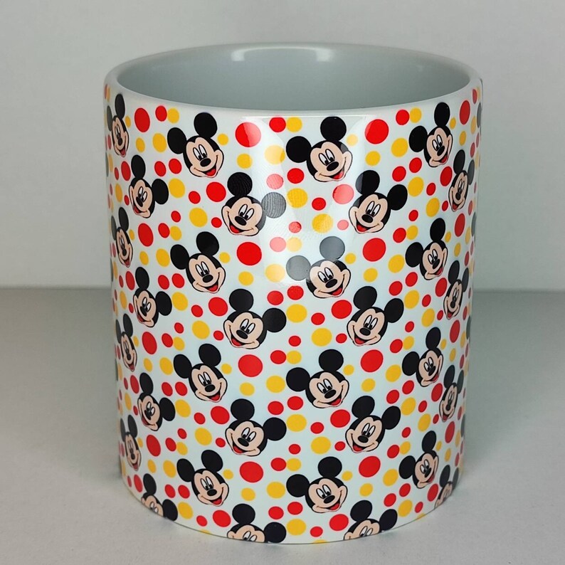 K&ouml;nnte beinhalten: Wei&szlig;e Keramiktasse mit einem sich wiederholenden Muster aus Mickey-Maus-K&ouml;pfen und gelben und roten Punkten.