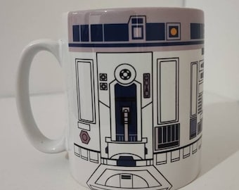 May include: White ceramic mug with a black and white illustration of R2-D2, a robot from the Star Wars franchise. The mug has a handle and a rounded shape.