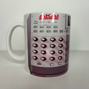 May include: White ceramic mug featuring a graphic of an Akai professional sampler. The graphic depicts a control panel with red knobs and buttons.