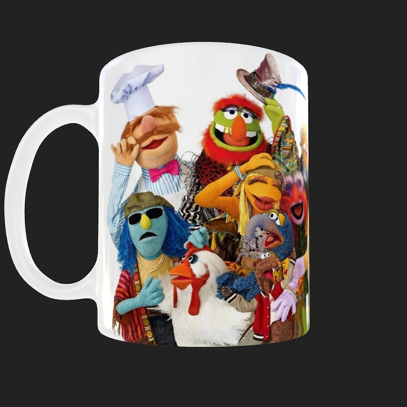 K&ouml;nnte beinhalten: Eine wei&szlig;e Keramiktasse mit einem bunten Bild der Muppet-Charaktere. Zu den Charakteren geh&ouml;ren Cookie Monster, Big Bird, Animal, Miss Piggy und andere. Die Tasse hat einen Henkel auf der linken Seite.