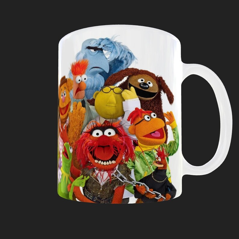 K&ouml;nnte beinhalten: Wei&szlig;e Keramiktasse mit einem bunten Bild von Sesamstra&szlig;e-Charakteren, darunter Big Bird, Elmo, Cookie Monster und Animal. Die Figuren sind in einer Gruppe angeordnet, wobei Animal im Vordergrund steht.