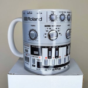 Puede incluir: Una taza de cerámica blanca con un diseño de caja de ritmos Roland TR-808. La taza presenta una imagen en blanco y negro del panel frontal de la caja de ritmos, incluidos los botones, botones y la pantalla icónicos. La taza tiene un asa blanca y un logotipo Roland negro.