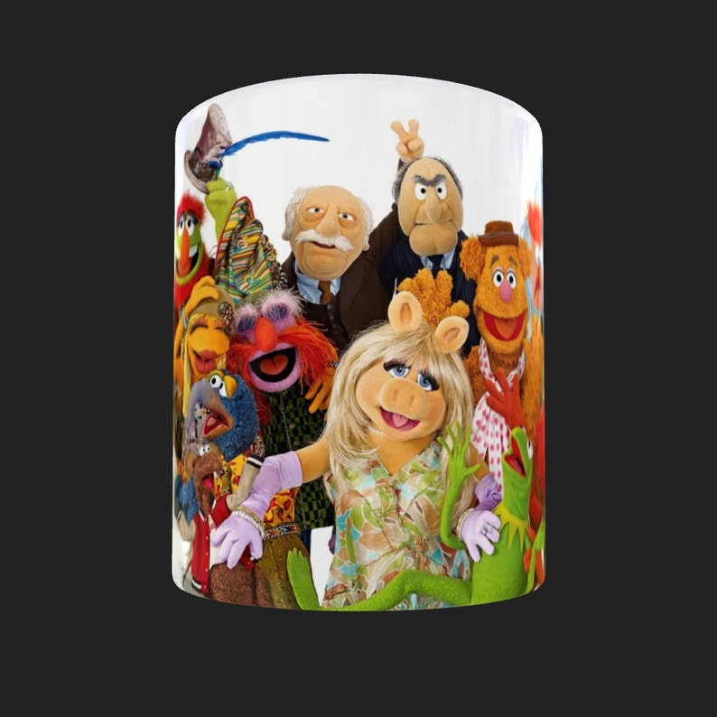 K&ouml;nnte beinhalten: Eine wei&szlig;e Keramiktasse mit einem bunten Bild der Muppet-Charaktere. Die Charaktere sind Miss Piggy, Kermit der Frosch, Fozzie B&auml;r, Gonzo, Animal und Scooter.