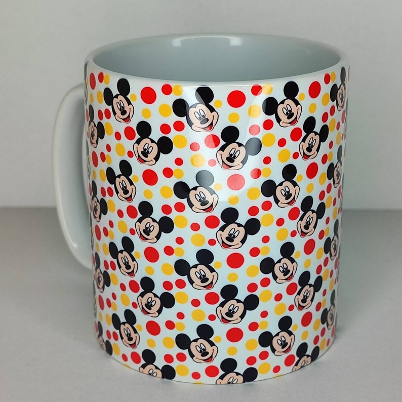 K&ouml;nnte beinhalten: Wei&szlig;e Keramiktasse mit einem sich wiederholenden Muster aus Mickey-Maus-K&ouml;pfen und roten und gelben Punkten.