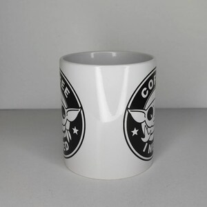 Pu&ograve; includere: Tazza da caff&egrave; in ceramica bianca con una grafica in bianco e nero di un personaggio dei cartoni animati con il testo "Coffee, I need it" in un cerchio.