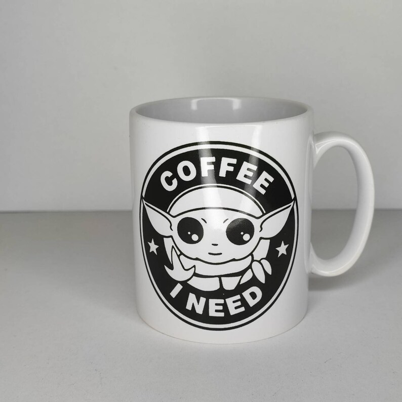 Pu&ograve; includere: Tazza da caff&egrave; in ceramica bianca con una grafica in bianco e nero di un personaggio dei cartoni animati con il testo "Ho bisogno di caff&egrave;" in un cerchio.