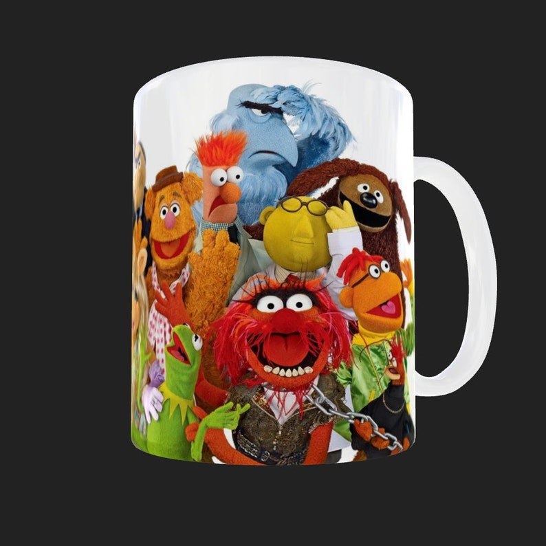 K&ouml;nnte beinhalten: Wei&szlig;e Keramiktasse mit einem bunten Bild der Muppet-Charaktere, darunter Animal, Cookie Monster und Miss Piggy.