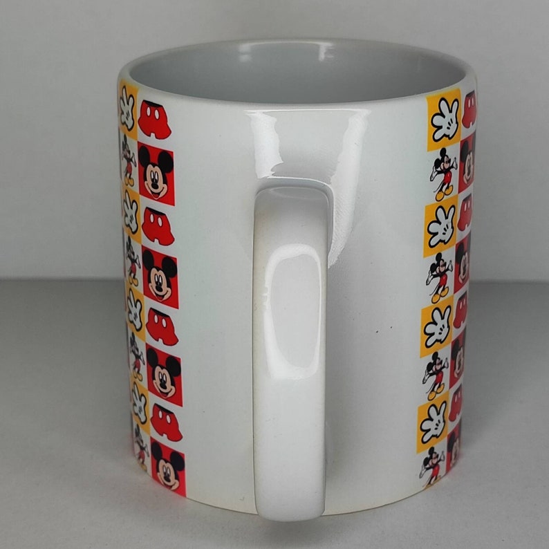 K&ouml;nnte beinhalten: Wei&szlig;e Keramiktasse mit einem schwarzen und roten Mickey Mouse Design. Die Tasse hat einen wei&szlig;en Henkel und ist mit einem sich wiederholenden Muster von Mickey Mouse Kopf, Handschuhen und roten Shorts verziert.