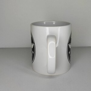 Pu&ograve; includere: Tazza in ceramica bianca con un logo in bianco e nero che presenta un aereo e stelle. La tazza ha un manico.