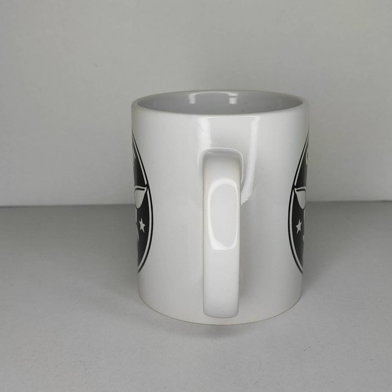 Pu&ograve; includere: Tazza in ceramica bianca con un logo in bianco e nero che presenta un aereo e stelle. La tazza ha un manico.