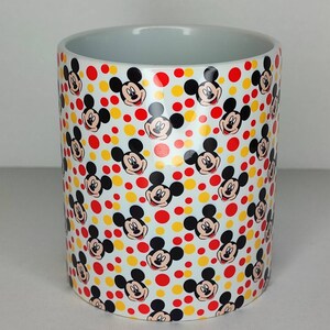 K&ouml;nnte beinhalten: Wei&szlig;e Keramiktasse mit einem sich wiederholenden Muster aus Mickey-Maus-K&ouml;pfen und gelben und roten Punkten.