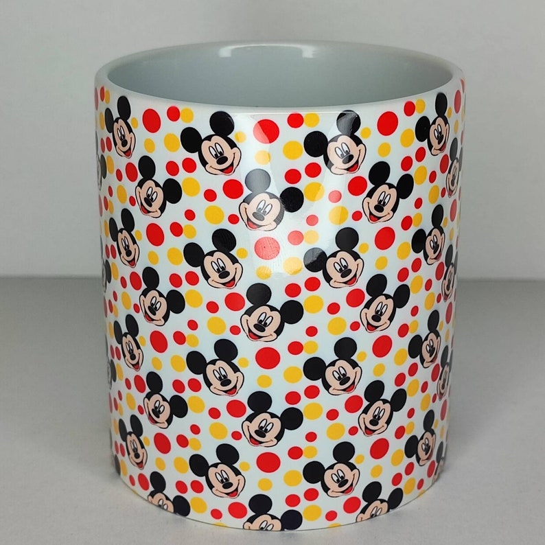 K&ouml;nnte beinhalten: Wei&szlig;e Keramiktasse mit einem sich wiederholenden Muster aus Mickey-Maus-K&ouml;pfen und gelben und roten Punkten.
