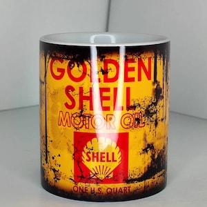 May include: A cylindrical mug with a vintage Golden Shell Motor Oil design. The mug has a yellow background with black distressed accents and red text. The Shell logo is displayed. The mug is labeled "ONE U.S. QUART."
