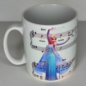 Könnte beinhalten: Weiße Keramiktasse mit einem aufgedruckten Bild eines Zeichentrickcharakters, Elsa aus Frozen, die auf einem Notenblatt singt. Der Text auf dem Notenblatt lautet "Em snow white on the king-dom of".