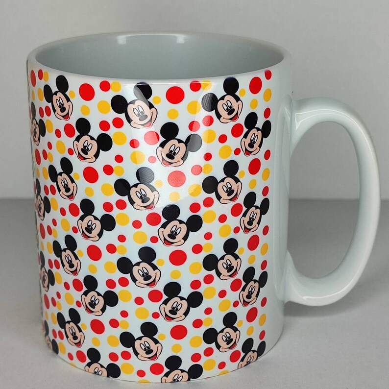 K&ouml;nnte beinhalten: Wei&szlig;e Keramiktasse mit einem sich wiederholenden Muster aus Mickey-Maus-K&ouml;pfen in Schwarz und Rot auf wei&szlig;em Hintergrund mit gelben und roten Punkten.