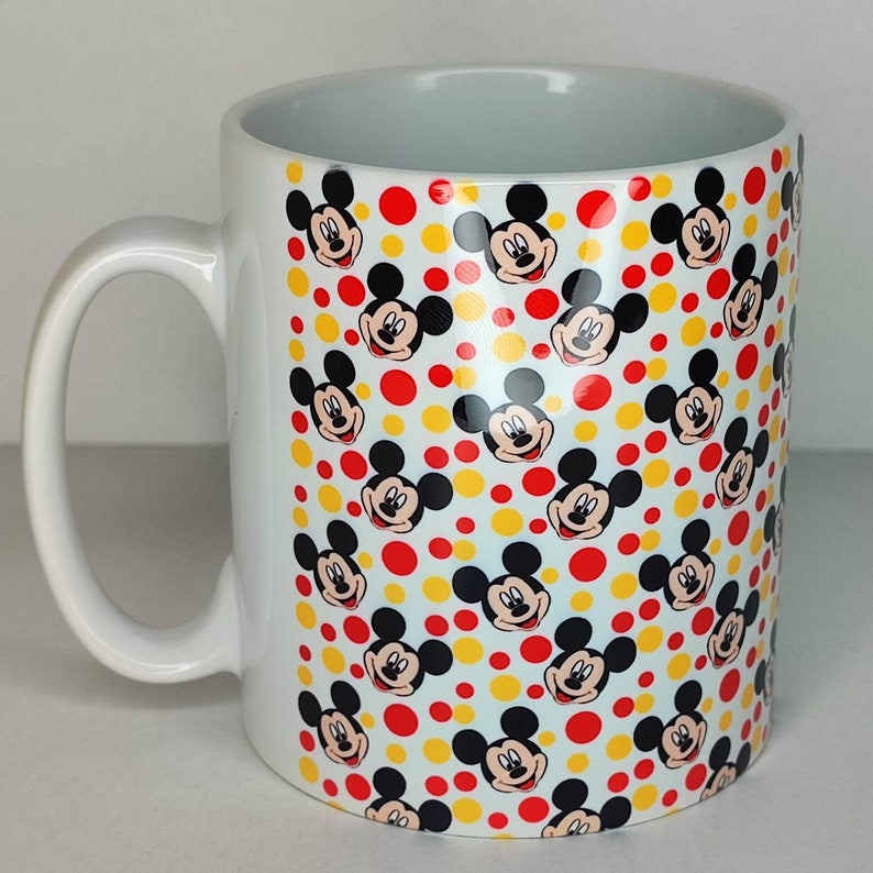 K&ouml;nnte beinhalten: Wei&szlig;e Keramiktasse mit einem sich wiederholenden Muster aus Mickey-Maus-Gesichtern und roten und gelben Punkten.