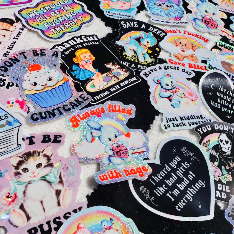 K&ouml;nnte beinhalten: Eine Sammlung farbenfroher, glitzernder Aufkleber mit verschiedenen Designs und Texten. Designs umfassen einen Cupcake, ein H&auml;schen mit Kamera, eine Katze und S&auml;tze wie "We should all be in therapy" und "I heard you like bad girls."