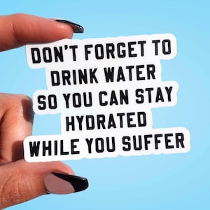Può includere: Un adesivo bianco con testo nero che recita: "NON DIMENTICARE DI BERE ACQUA COSÌ PUOI RIMANERE IDRATATO MENTRE SOFFRI". L'adesivo è tenuto tra le dita su uno sfondo azzurro.