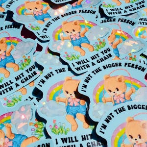 May include: A pile of light blue stickers. Each sticker features a cartoon teddy bear in blue overalls with a jump rope, a rainbow, and the text "I WILL HIT YOU WITH A CHAIR" and "I'M NOT THE BIGGER PERSON". The stickers have a holographic effect.