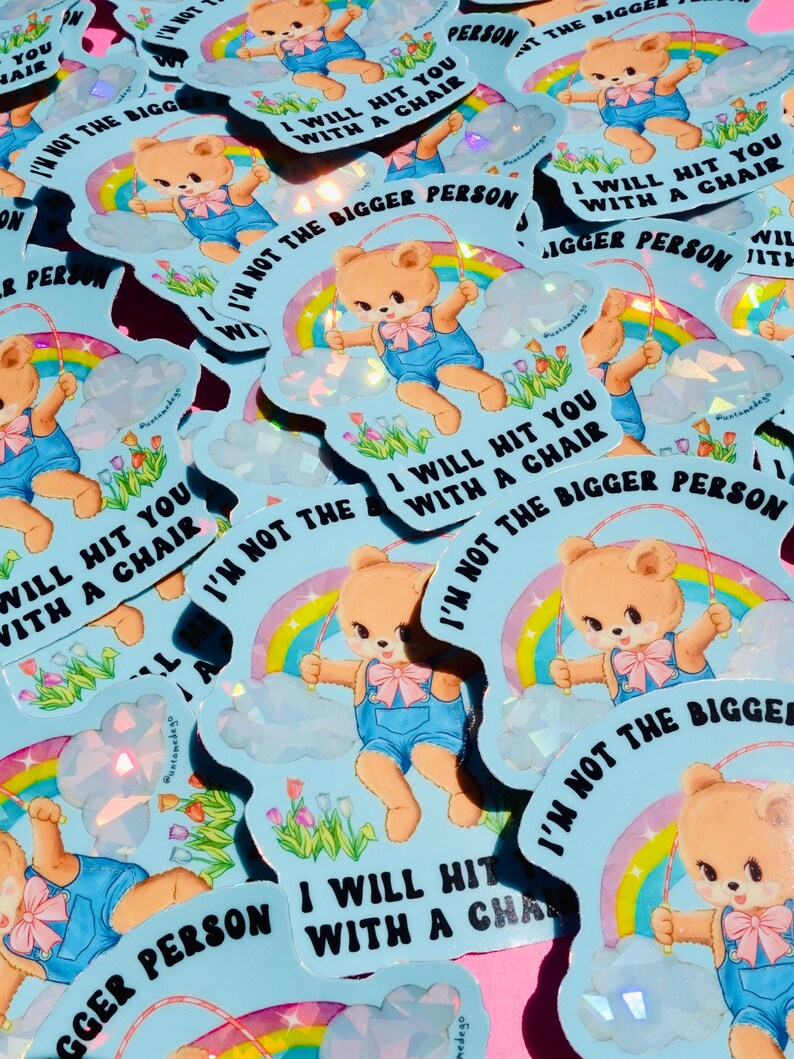 May include: A pile of light blue stickers. Each sticker features a cartoon teddy bear in blue overalls with a jump rope, a rainbow, and the text "I WILL HIT YOU WITH A CHAIR" and "I'M NOT THE BIGGER PERSON". The stickers have a holographic effect.