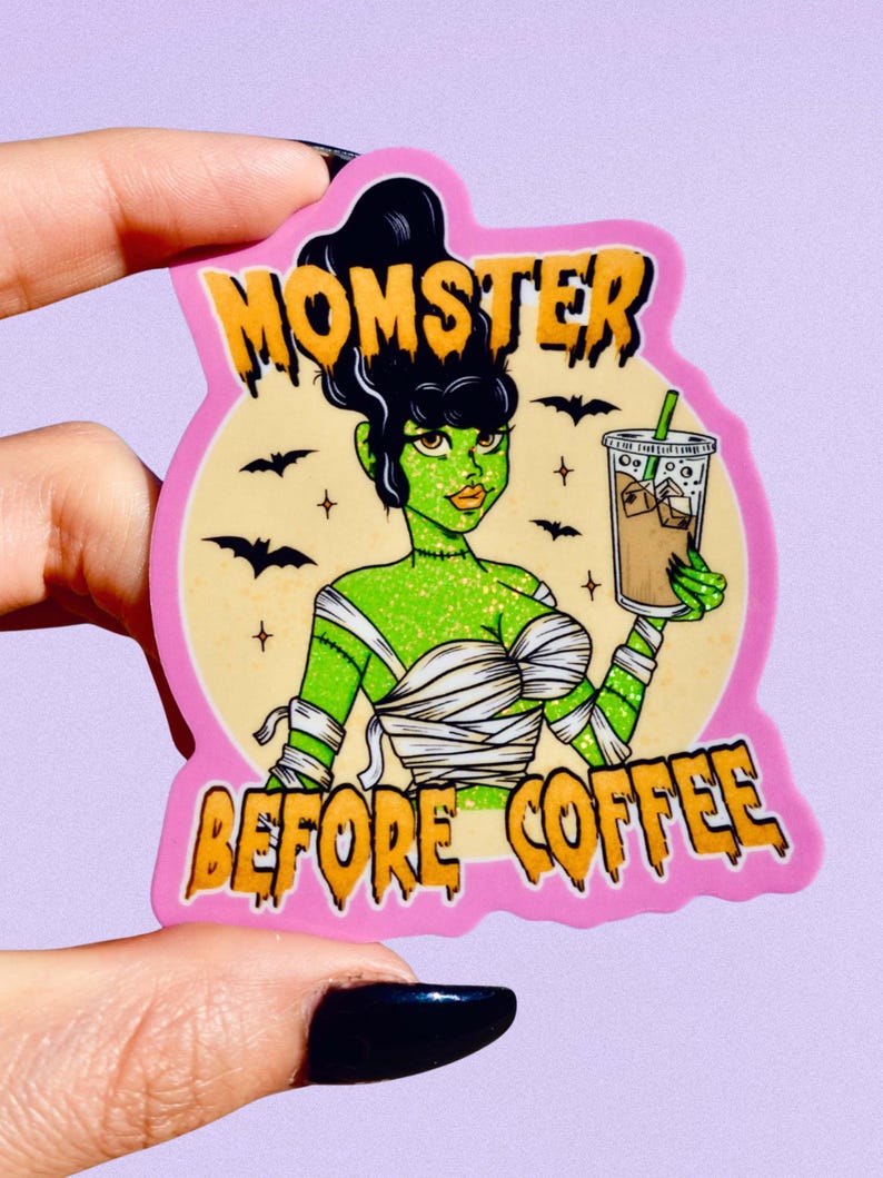 May include: A pink sticker with the words "MOMSTER BEFORE COFFEE" in orange lettering. The sticker features a green-skinned monster with black hair, wrapped in bandages, holding an iced coffee drink. Black bats and stars are in the background.