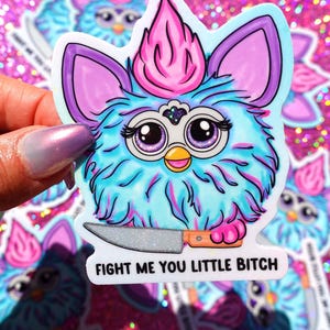 Puede incluir: Una pegatina con un personaje de dibujos animados parecido a un Furby con pelaje azul y morado, ojos grandes y una cresta rosa. El personaje sostiene un cuchillo, con el texto "FIGHT ME YOU LITTLE BITCH" debajo.