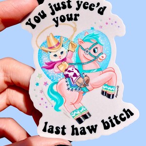 Op de afbeelding: Een sticker met een cartoonkat met een cowboyhoed die op een roze paard rijdt. Het paard heeft turquoise haar en accenten. De sticker heeft de tekst "You just yee'd your last haw bitch".