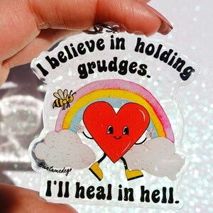 May include: A clear acrylic keychain with a rainbow, a heart with a face, and a bee. The text on the keychain reads "I believe in holding grudges. I'll heal in hell."