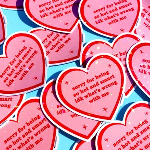 Pu&ograve; includere: Una collezione di adesivi rosa a forma di cuore con bordo rosso e la scritta "sorry for being so hot and smart idk what's wrong with me". Gli adesivi hanno un effetto glitterato e sono sparsi su una superficie azzurra.