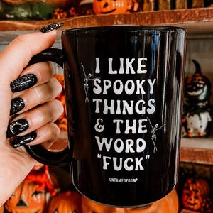 May include: A black ceramic mug featuring white text that reads I LIKE SPOOKY THINGS & THE WORD FUCK with small white skeleton illustrations on the sides and the brand name UNTAMEDEGO with a heart symbol printed at the bottom center.