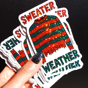 May include: A stack of stickers featuring a red and green striped sweater design with the words "Sweater Weather" in a dripping font. The stickers have a glittery effect and are held against a black background.