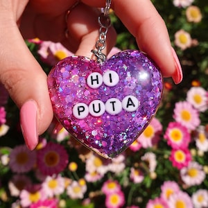 May include: A pink and purple heart-shaped keychain with glitter embedded in the resin. The keychain has white letters spelling out "HI PUTA".