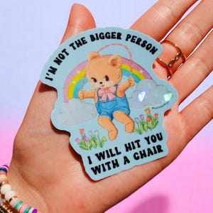 May include: A light blue sticker featuring a cartoon bear skipping rope in front of a rainbow. The text on the sticker reads "I'M NOT THE BIGGER PERSON I WILL HIT YOU WITH A CHAIR."