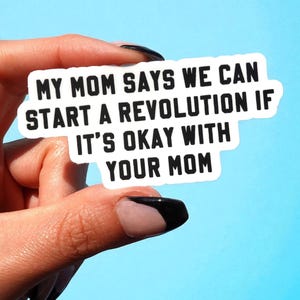 May include: A white rectangular sticker with black text that reads: "MY MOM SAYS WE CAN START A REVOLUTION IF IT'S OKAY WITH YOUR MOM." The sticker is held against a light blue background.